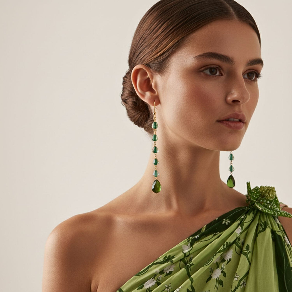 A woman with smooth brown hair in a low bun wears the Holly Quartz Dangle Hook Earrings by JAREDJAMIN Jewelry Online. Dressed in a green, floral one-shoulder dress, she looks slightly to the side against a plain light background.