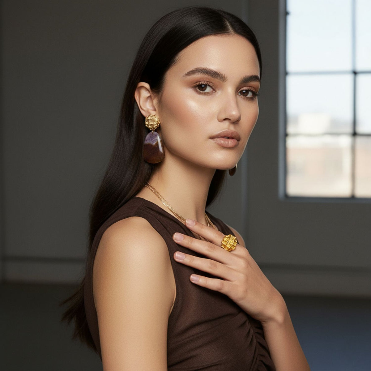 A woman with long dark hair poses by a window, wearing a sleeveless brown dress, gold ring, necklace, and JAREDJAMIN Jewelry Online’s Honeycomb Clip on Earrings with Jasper. Her hand rests on her chest as she gazes softly at the camera.