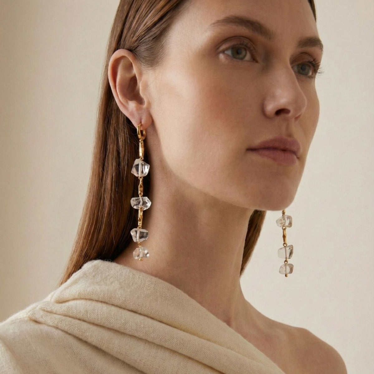 Un woman avec straight brown hair et light skin models the Ice Dangle or Leverback boucles d'oreilles par JAREDJAMIN Bijoux en Ligne—18K plaqué or, embellished avec clear geometric crystals. She wears a cream wrap et gazes upward against a beige background.