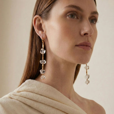 A woman with straight brown hair and light skin models the Ice Dangle Gold Leverback Earrings by JAREDJAMIN Jewelry Online—18K gold-plated, embellished with clear geometric crystals. She wears a cream wrap and gazes upward against a beige background.