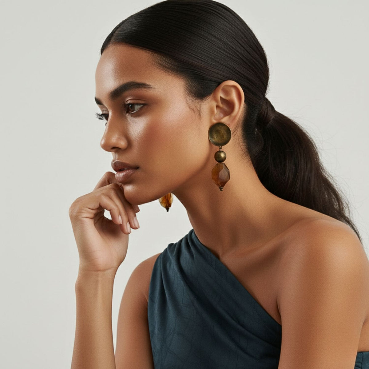 A woman with smooth, dark hair in a low ponytail gazes to the left, resting her chin on her hand. She wears JAREDJAMIN Jewelry Online’s Iron Bowl Clip Earrings and a dark, one-shoulder top, set against a plain light background.