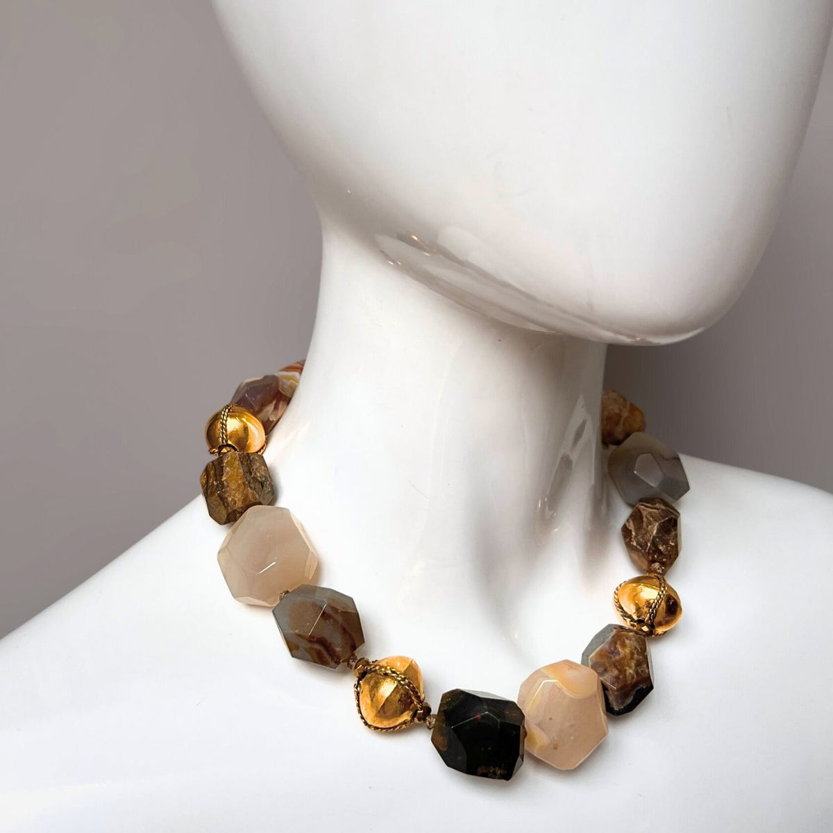 In a close-up shot, the Isolde Agate Necklace by JAREDJAMIN Jewelry Online is draped on a mannequin, displaying chunky agate gemstones in brown, cream, and amber. Geometric stones intermixed with textured 18k gold plate beads create a striking design against a gray backdrop.