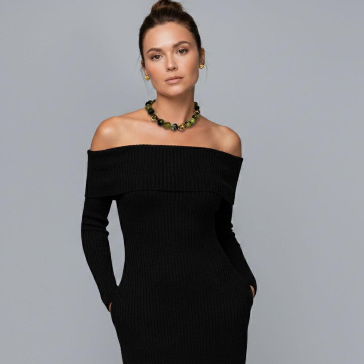 A woman with brown hair in a bun wears a black off-the-shoulder ribbed dress and the Moss Maiden Necklace by JAREDJAMIN Jewelry Online. She stands confidently with hands in her pockets against a light gray background, looking at the camera.