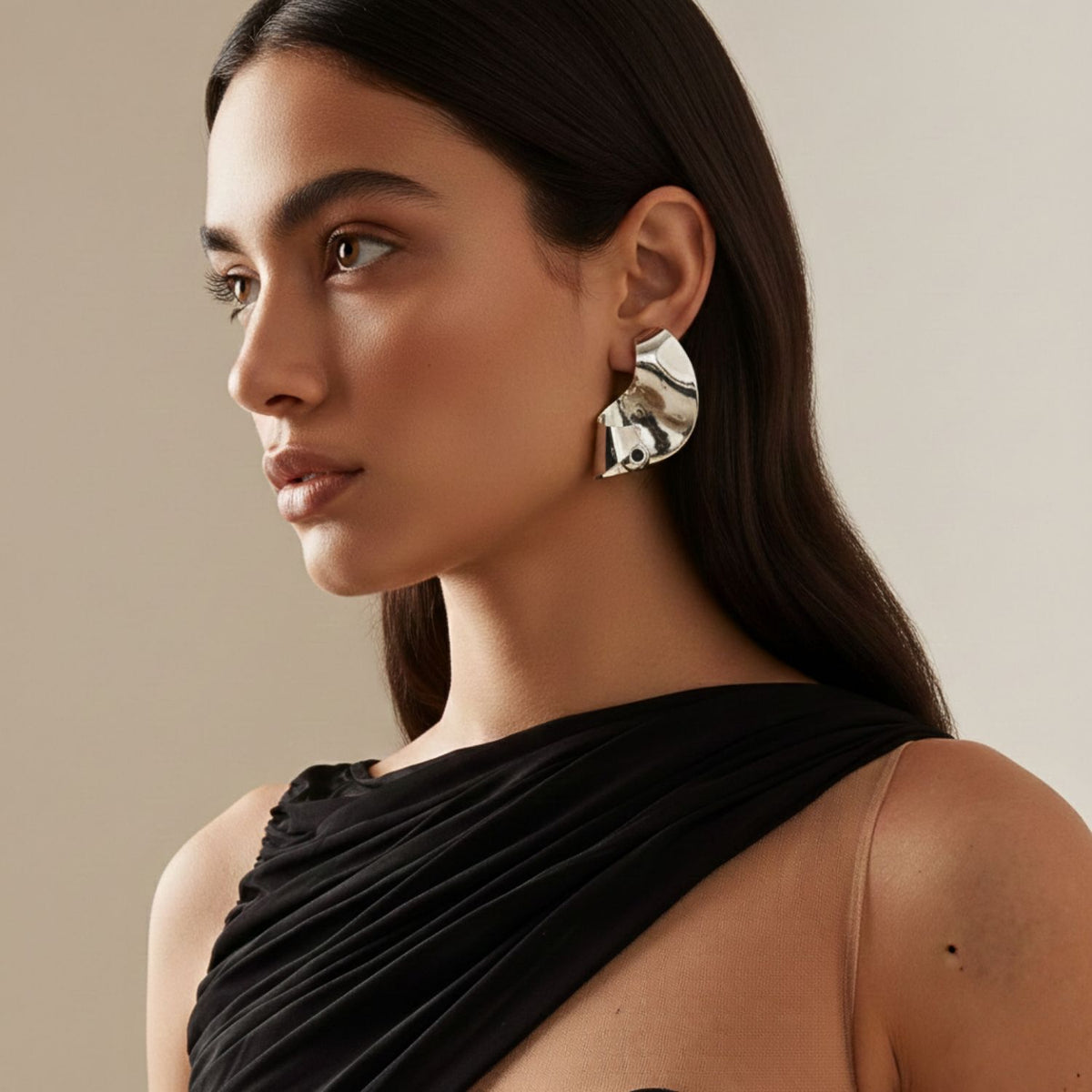A womanとlong, dark hair wears a one-shoulder ブラック dressとOnda Ball イヤリング JAREDJAMIN製 ジュエリーオンライン. She gazes sideways 付きの neutral expression against a plain beige background, her makeup naturalとsubtle.