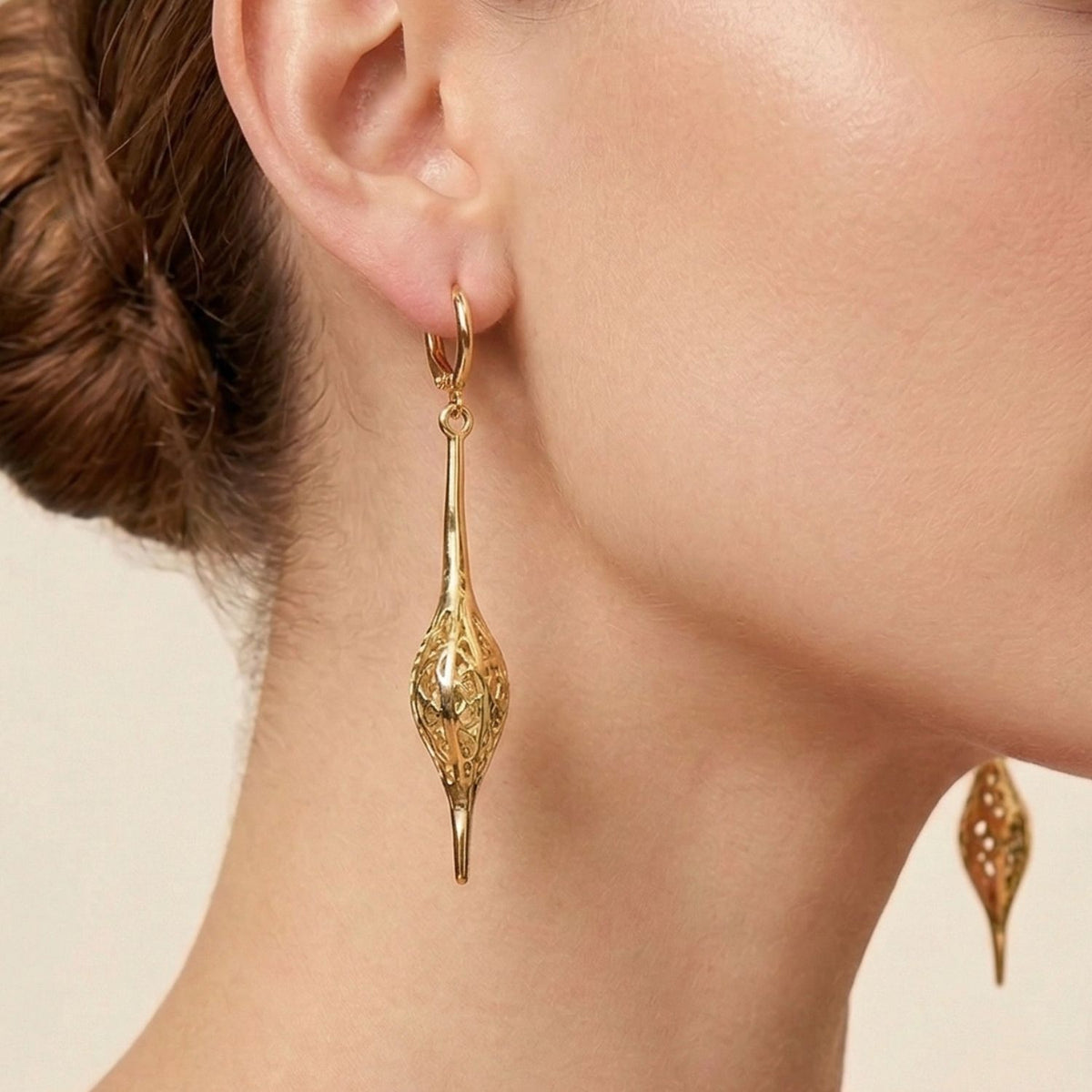 Close-up of a woman's ear wearing Jeannie Drop Lever Back Earrings from JAREDJAMIN Jewelry Online, featuring a long 18k gold-plated dangle with intricate filigree. Her hair is in a low bun, highlighting her neck and jawline against a neutral background.