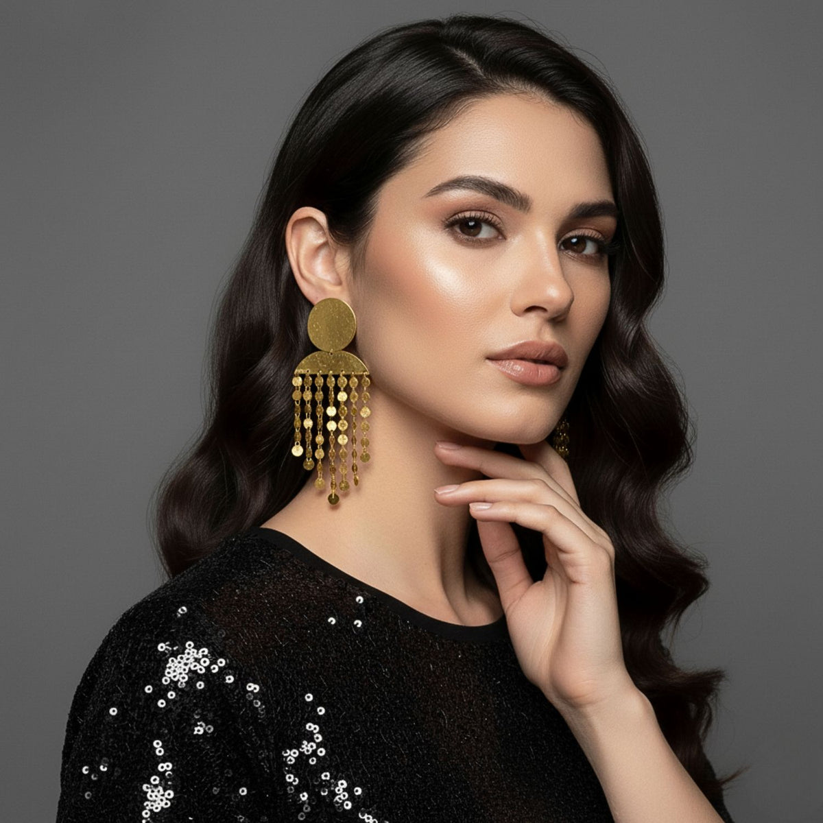 A woman with wavy dark brown hair, in a black sequin top, poses against a gray background. She wears Koren Clip On Earrings from JAREDJAMIN Jewelry Online—large, round 18k gold statement earrings with dangling chains.