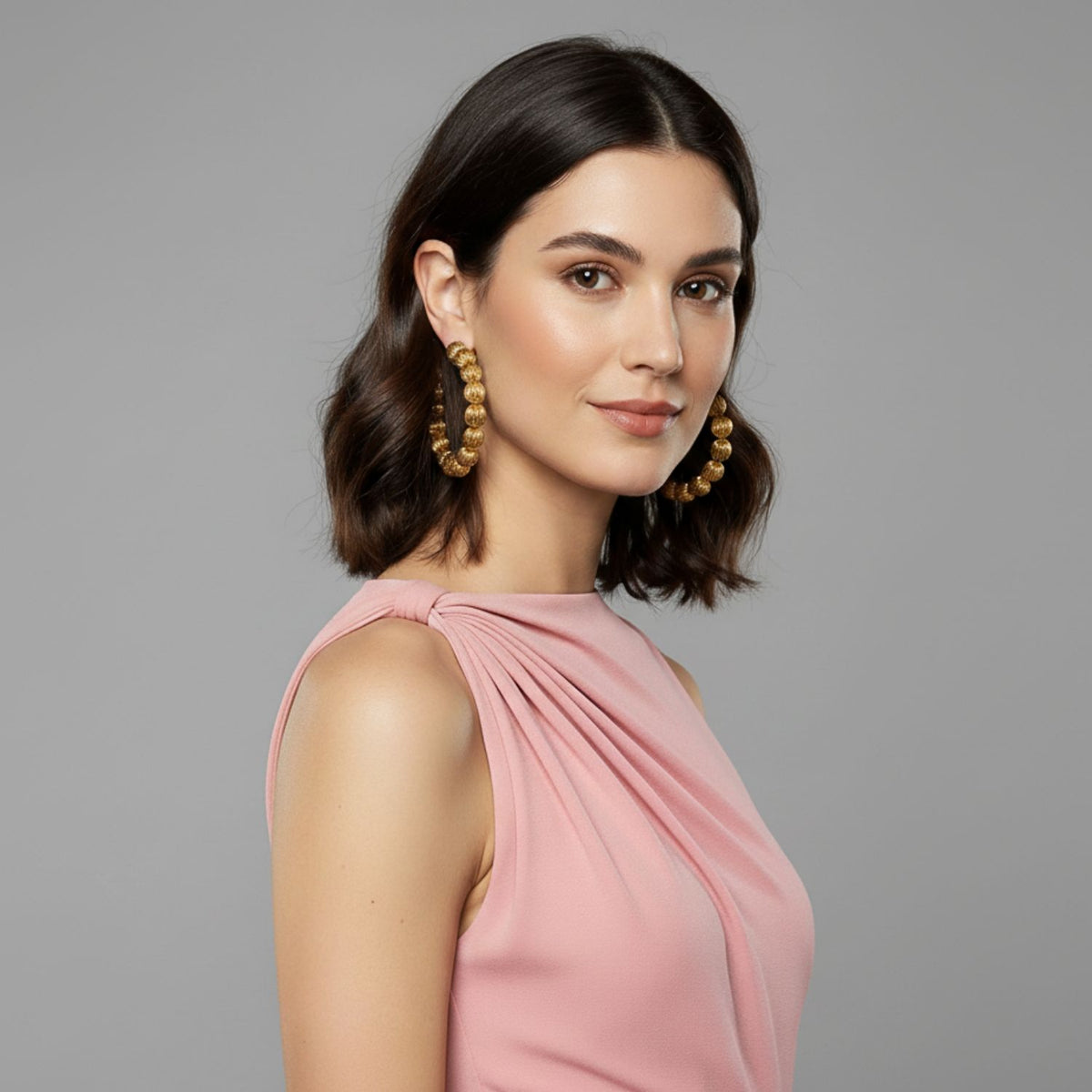 Un woman avec shoulder-length brown hair in loose waves wears a sleeveless pink dress et Large or Lantern créole Post boucles d'oreilles par JAREDJAMIN Bijoux en Ligne. She poses avec natural makeup against a plain light gray background.