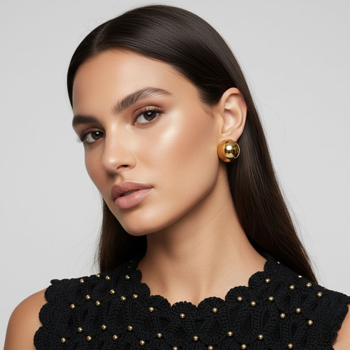 A woman with long, straight dark hair and glowing makeup gazes at the camera. She wears a sleeveless black top with gold studs and JAREDJAMIN Jewelry Online's Gold Large Orbita Clip Earrings, making a bold statement on a light gray background.