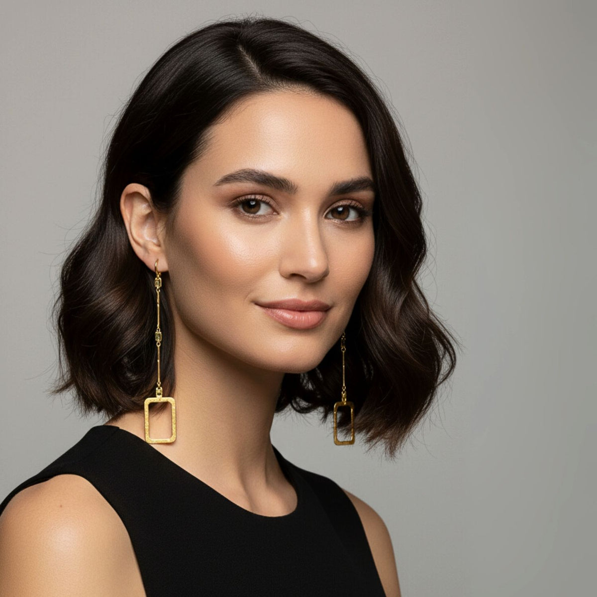 A woman with wavy, shoulder-length dark hair wears a sleeveless black top and the Lattice of Gladys Drop Earrings by JAREDJAMIN Jewelry Online, finished in 18K gold plating. She faces forward, smiling softly with neutral makeup against a light gray background.