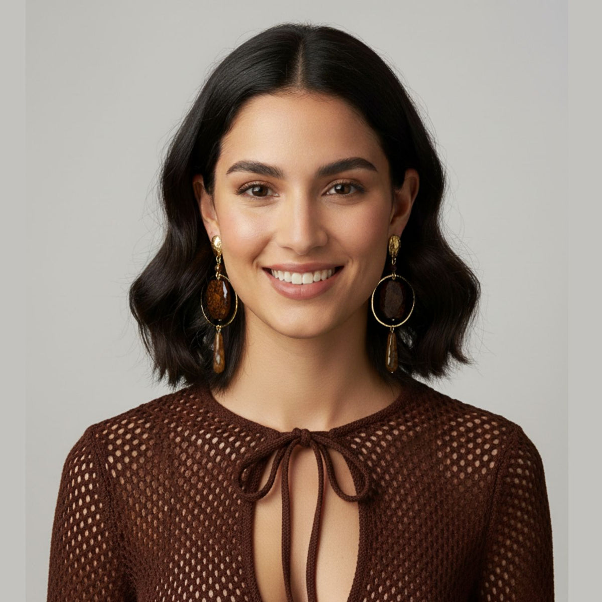 Un woman con wavy, shoulder-length dark hair smiles at the camera, wearing JAREDJAMIN Joyería Online’s Lindy Creek Agate pendientes y a brown long-sleeve mesh-knit top con a bow tie neckline. Los background is plain y light gray.