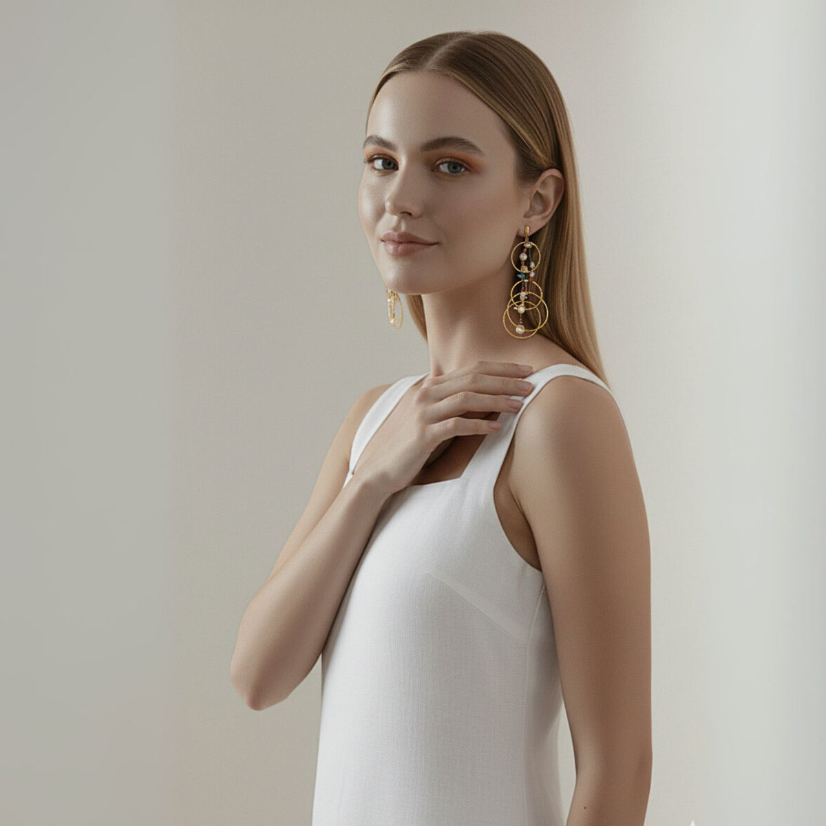A woman with straight, light brown hair wears a sleeveless white dress and elegant Lindy Dream Catcher Dangle Earrings by JAREDJAMIN Jewelry Online. She smiles gently at the camera, her right hand on her collarbone against a softly lit background.