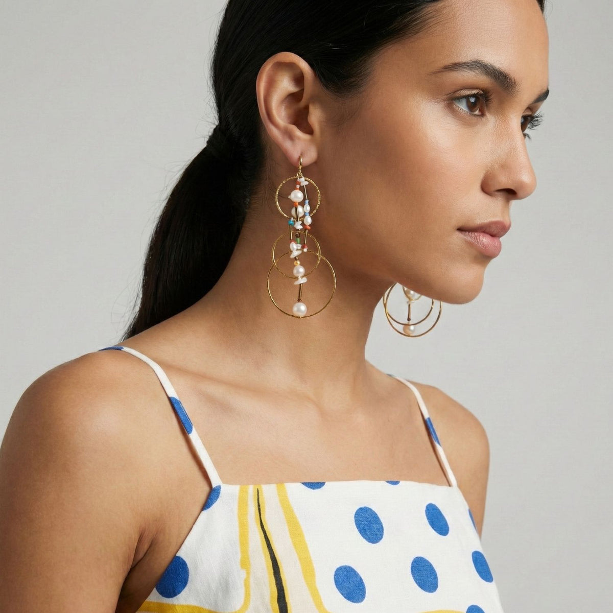 A woman with long dark hair in a low ponytail wears Lindy Dream Catcher Dangle Earrings by JAREDJAMIN Jewelry Online—18K gold with small beads and pearls. She pairs them with a white spaghetti-strap dress featuring blue dots and yellow patterns.