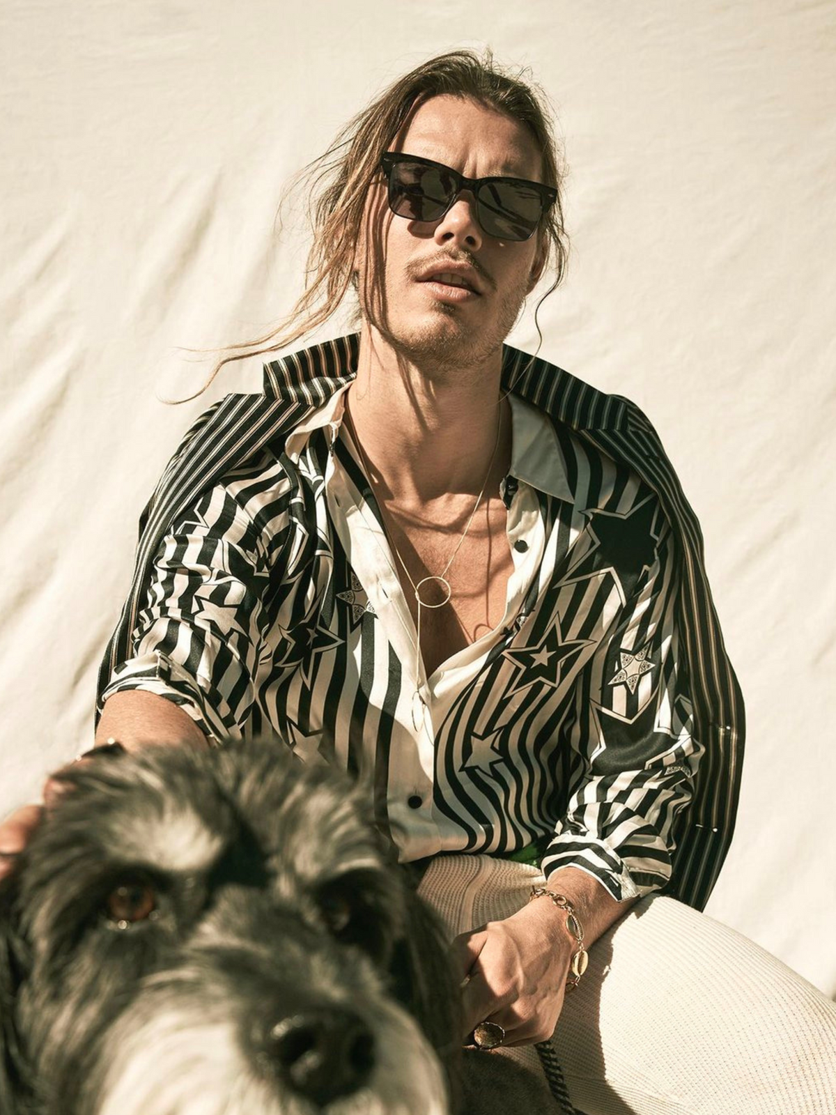 A person in sunglasses and a star-patterned striped shirt sits against a light background. Their shoulder-length hair complements the tarnish-resistant Lindy Y-Necklace from JAREDJAMIN Jewelry Online, while they gently rest their hand on the head of a large black and white dog.