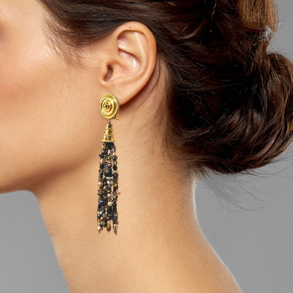 A woman with dark hair in a loose updo wears JAREDJAMIN Jewelry Online’s Loquacious Tassel Clip On Earring, an 18k gold plated circular piece with long black beaded tassels and gold accents. A plain gray background highlights her features.