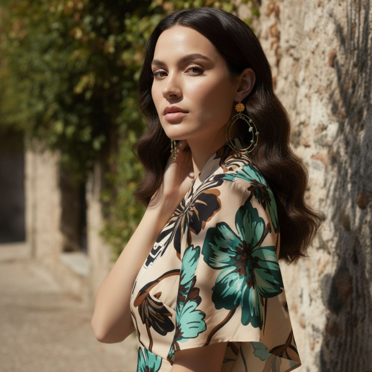 A womanとlong, wavy dark hair stands by a テクスチャード stone wall outdoors, wearing a beige dressとグリーンとbrown floralsとJAREDJAMIN ジュエリーオンライン’s Lotus Dream Aventurine Chandelier イヤリング. Sunlight highlights her confident gaze at camera.