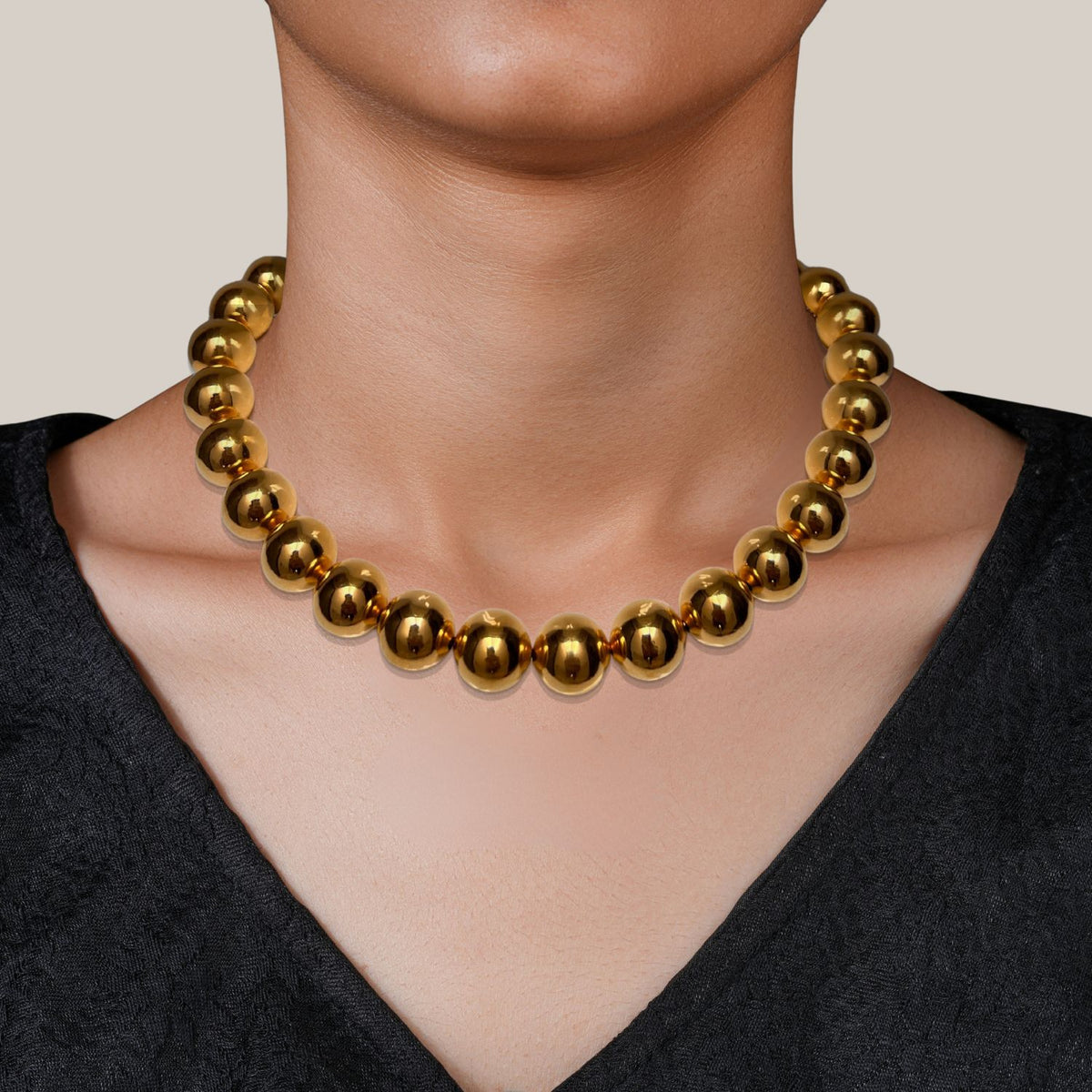 Wearing the Lucille Ball Necklace by JAREDJAMIN Jewelry Online—an 18k gold piece with bold, shiny beads—a person in a textured black top poses against a light beige background, showcasing only their lower face, neck, and collarbone.