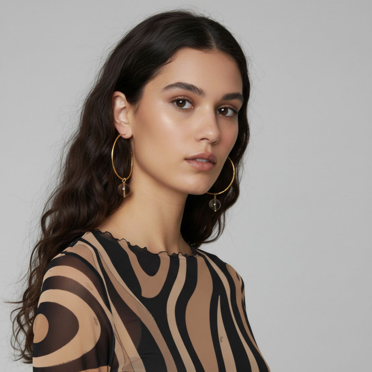 A woman with long, wavy dark hair wears JAREDJAMIN Jewelry Online’s Hooptastic Gemstone Drop Gold Hoop Earrings and a sheer patterned top with black and tan swirls. She faces left against a plain gray background, expression neutral and makeup natural.