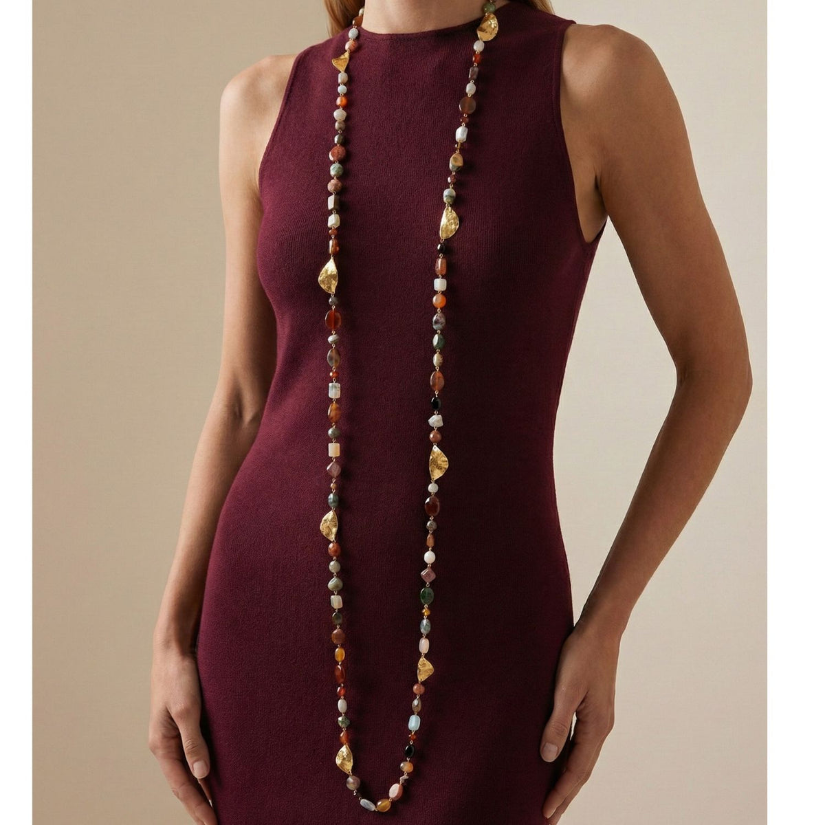 A woman in a sleeveless burgundy dress wears the Geo Twist Gemstone Linked Necklace by JAREDJAMIN Jewelry Online, featuring jasper agate quartz and gold-toned leaf accents. The necklace drapes to her waist against a neutral background.