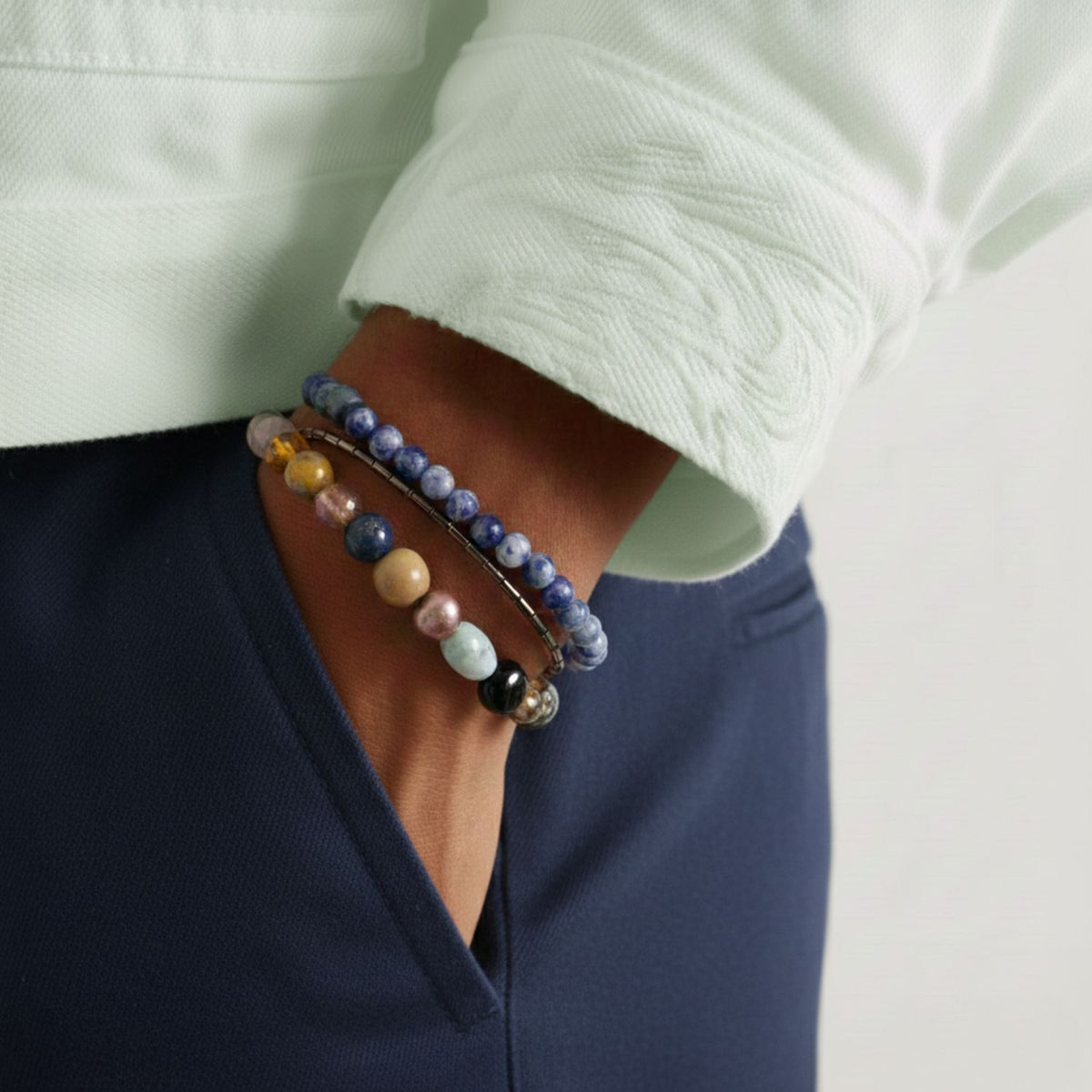 A person in a light mint green jacket and navy blue pants wears the Maurice Gemstone Bracelet Set by JAREDJAMIN Jewelry Online, featuring elastic bracelets with hematite, blue, yellow, black, and gemstone-glass blend beads.