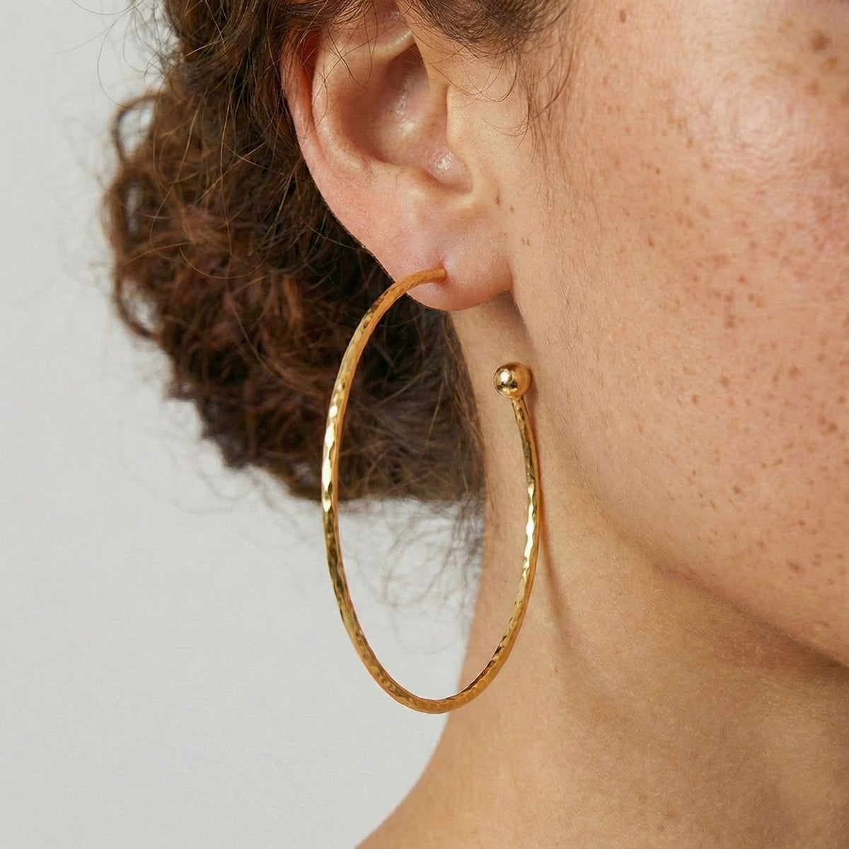 gros plan of a woman's ear wearing JAREDJAMIN Bijoux en Ligne's or Medium Hooptastic Post boucles d'oreilles, 18K or, shown against her light freckled skin et curly brown hair pour un standout, never-tarnish look.