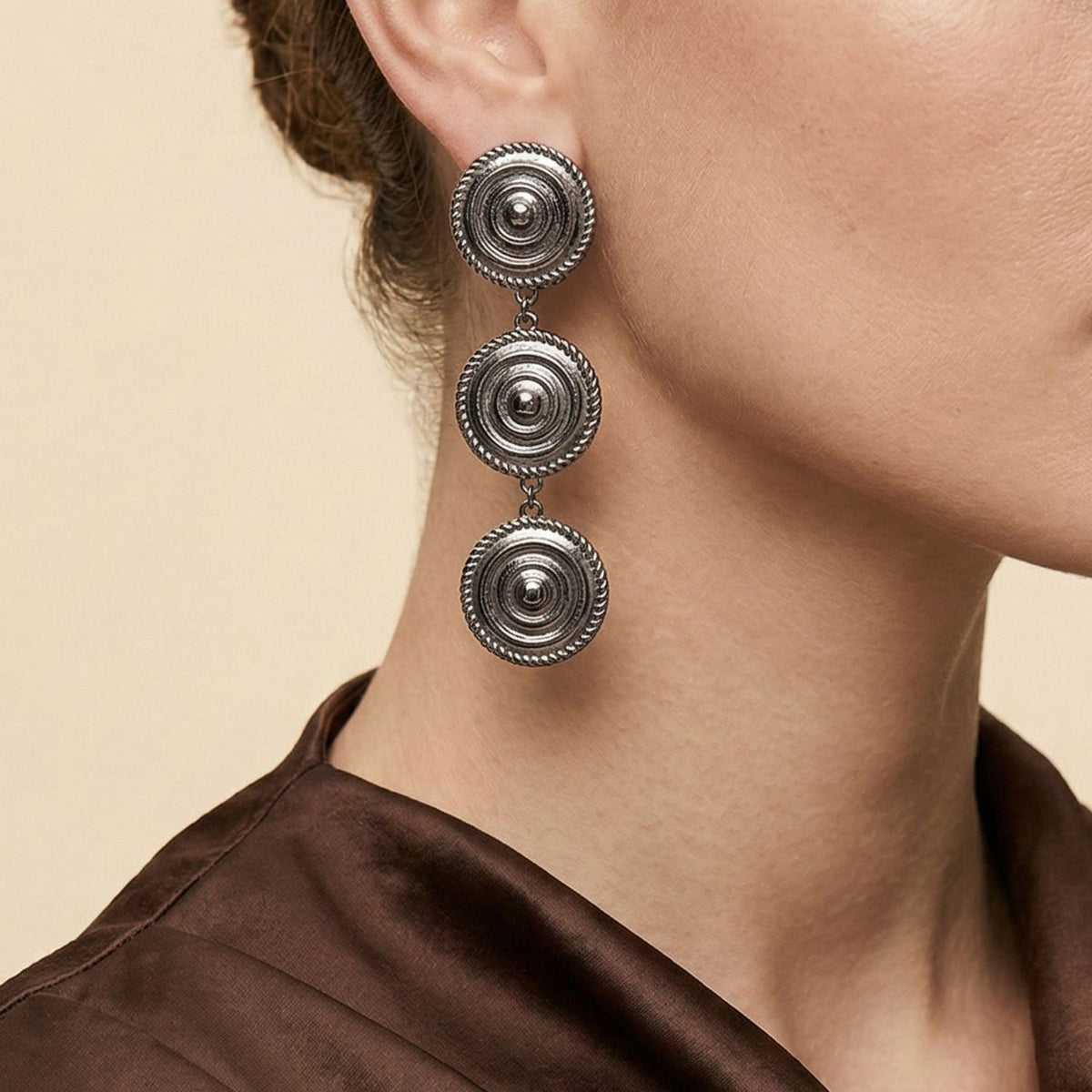 A woman with fair skin wears a brown satin top and Minerva Black Clip on Earrings by JAREDJAMIN Jewelry Online—large, silver, circular clip-ons with three stacked discs. Her brunette hair is pulled back against a plain beige background.