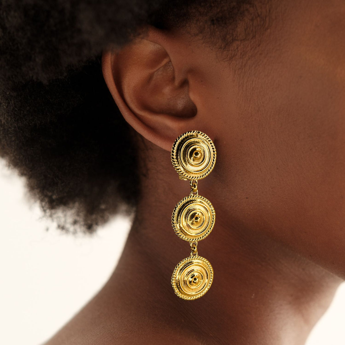 Close-up of a person wearing Minerva Gold Clip on Earrings by JAREDJAMIN Jewelry Online—bold 18K gold-plated dangles with three linked circles and textured edges, shown beside the ear against dark skin and curly hair.
