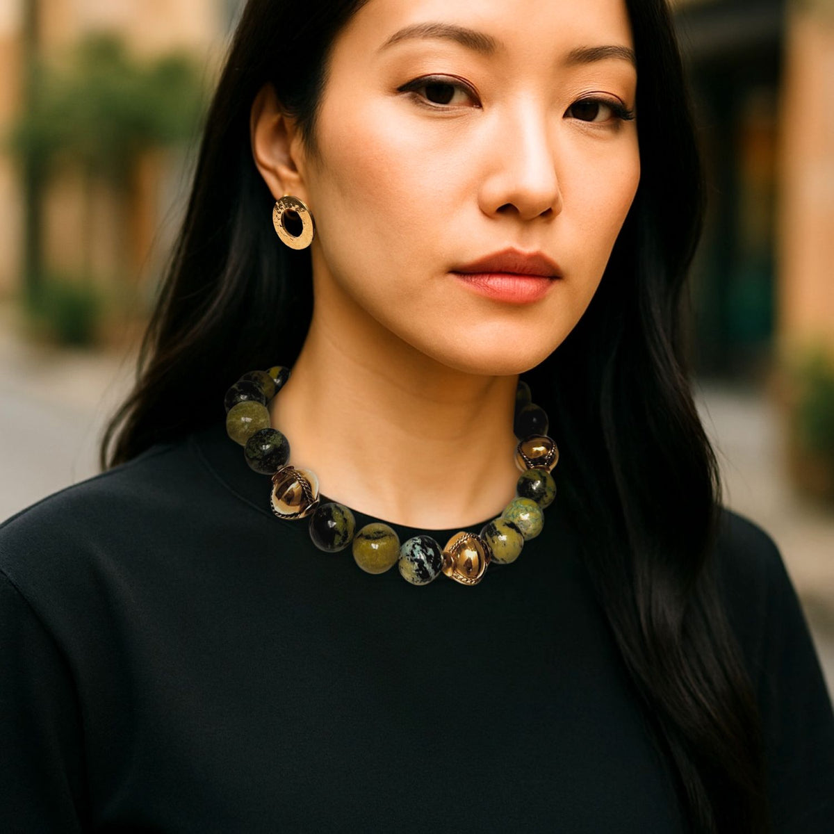 A woman with straight black hair wears a black top, 18K gold-plated hoops, and the Moss Maiden Necklace by JAREDJAMIN Jewelry Online. She stands outdoors, calmly facing the camera with a blurred street in the background.