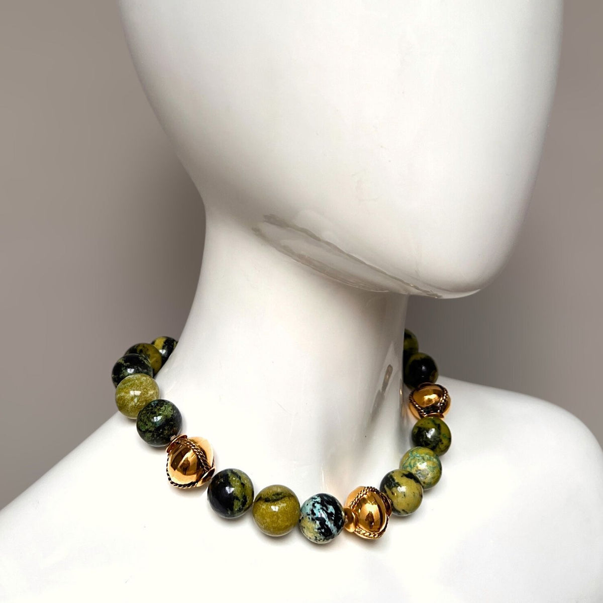 Close-up of a mannequin wearing the Moss Maiden Necklace by JAREDJAMIN Jewelry Online, featuring 18K gold-plated beads with green moss agate and brown marbled spheres. The mannequin’s smooth, glossy surface is set against a neutral backdrop.