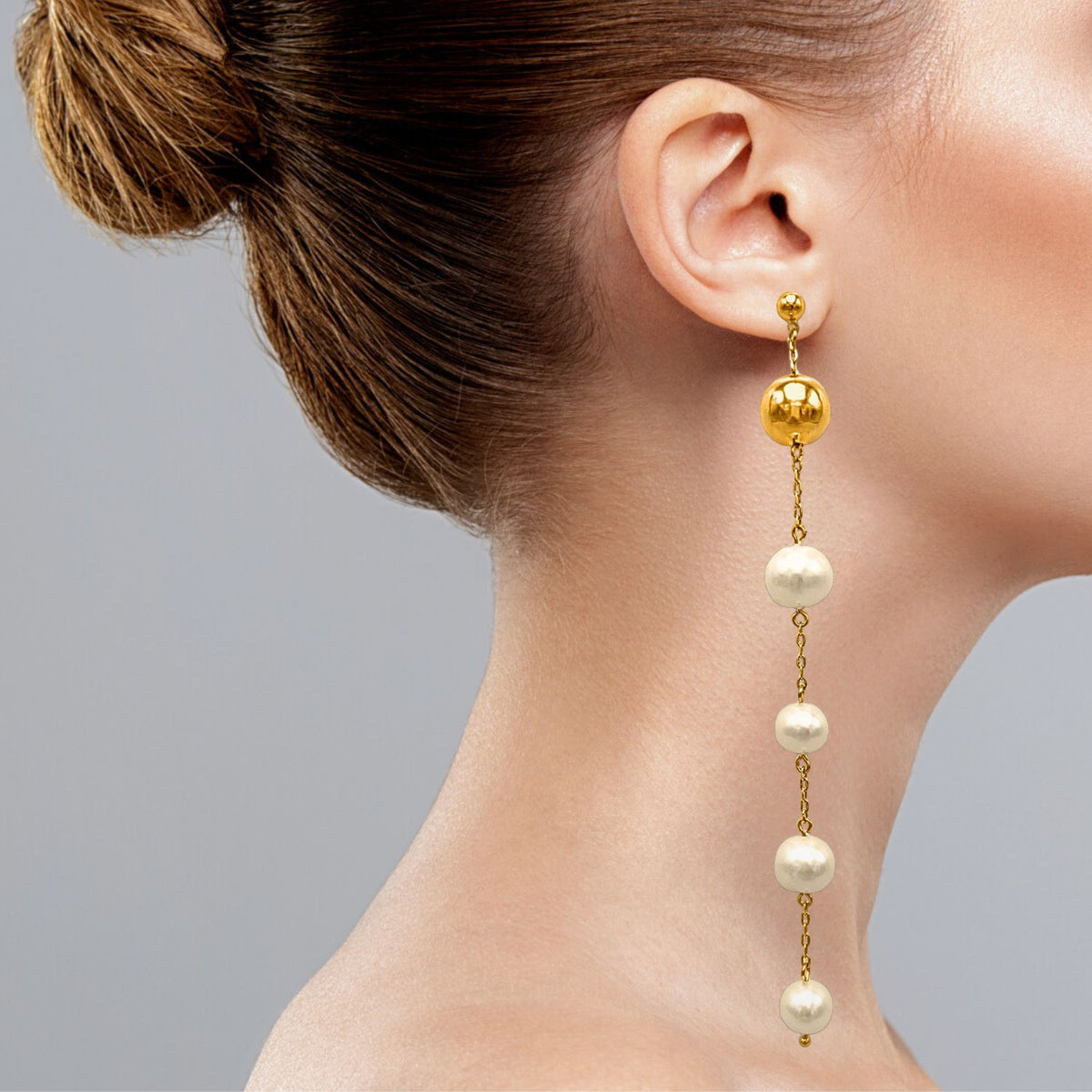 A woman with a bun hairstyle elegantly displays the Nora Dangle Pearl Earrings by JAREDJAMIN Jewelry Online, featuring an 18k gold stud and chain with four large pearls, set against a smooth light gray background that complements the refined jewelry and neat hairstyle.