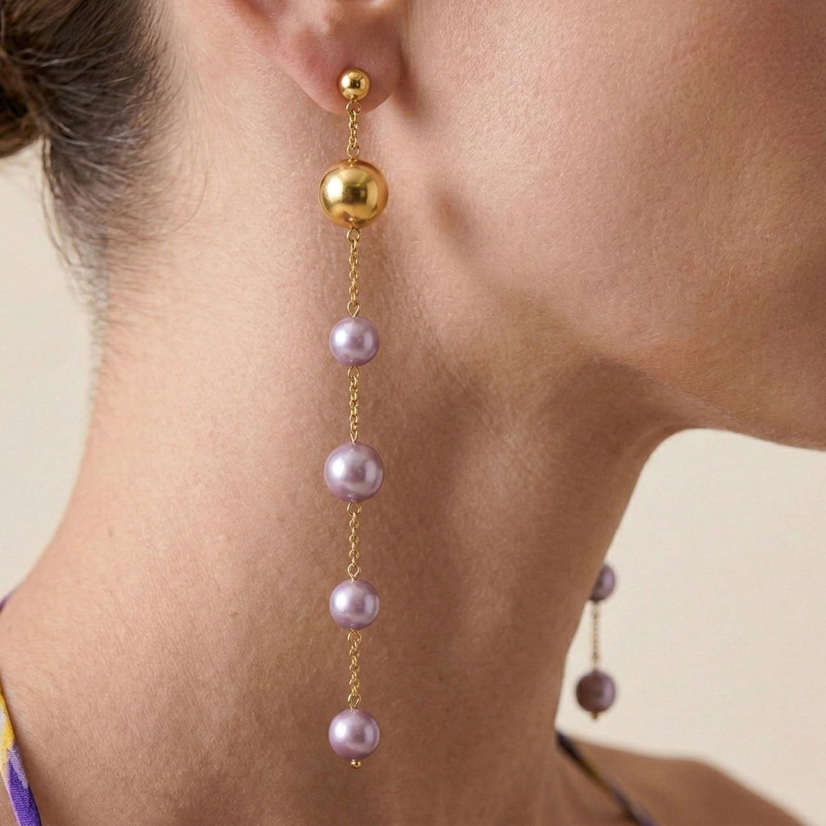 A close-up of a person's ear wearing the Nora Dangle Pearl Lavender Earrings by JAREDJAMIN Jewelry Online, featuring four spaced lavender freshwater pearls on an 18k gold-plated stud, with part of the neck and jawline shown.