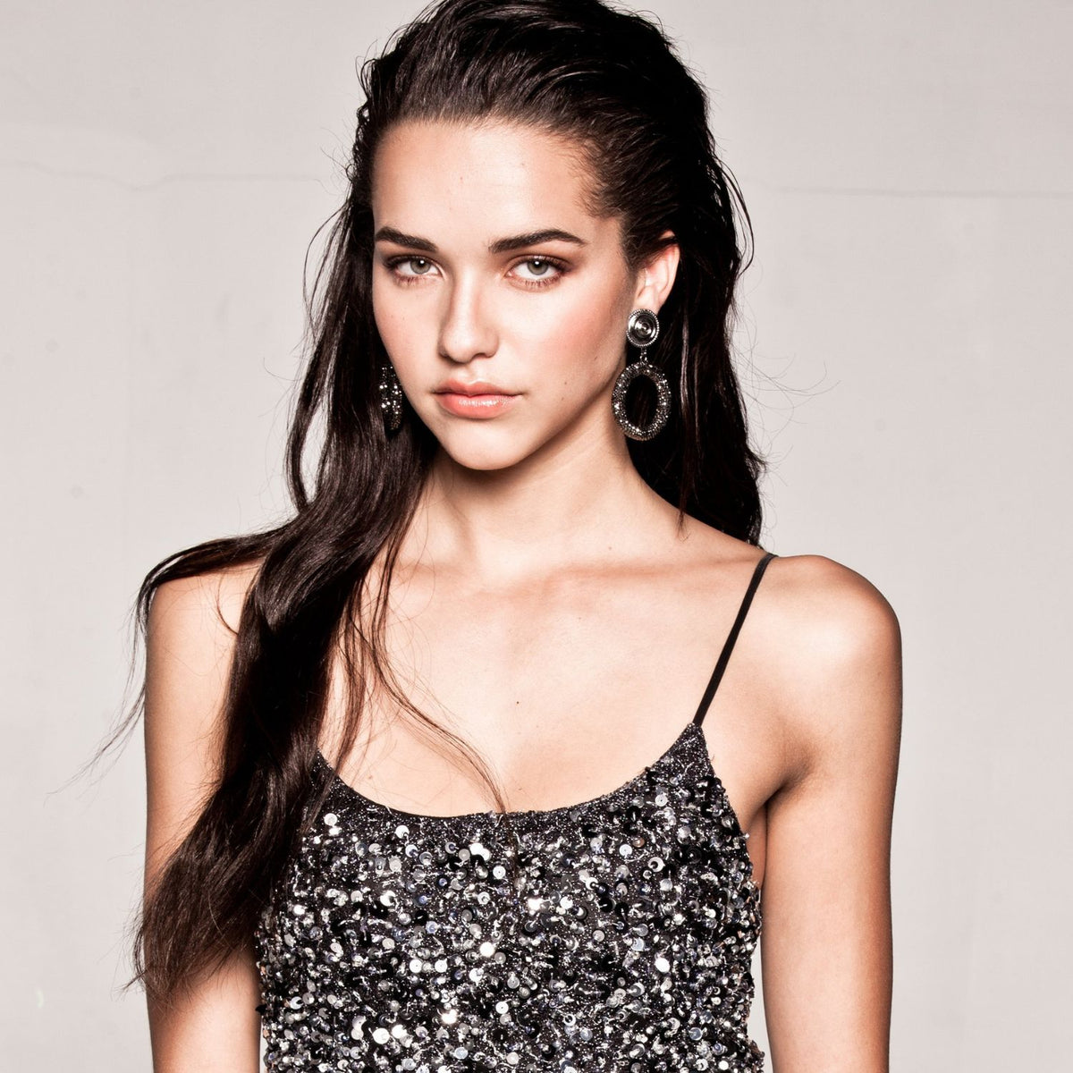 A young woman with long dark hair wears the Dolly Pavé Clip On Earrings by JAREDJAMIN Jewelry Online and a sequined spaghetti strap top. She stands against a neutral background with a confident expression, slightly tilted head, and natural makeup.