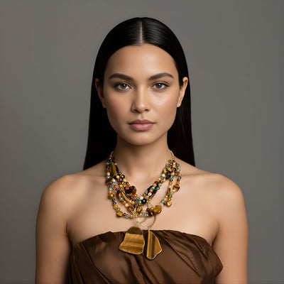 A woman with long, straight dark hair poses against a gray background, wearing a strapless brown top and the Sybil Tiger's Eye Gemstone Necklace from JAREDJAMIN Jewelry Online, featuring chunky gold, brown, and amber strands. Her expression is calm.