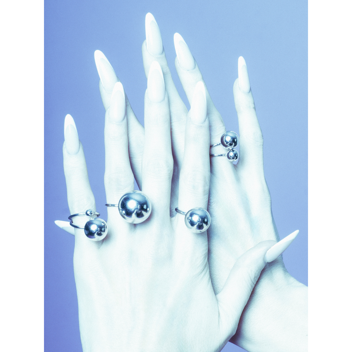 Two pale hands con long, white nails overlap, each adorned con the Orbita plata Three anillo Stack de JAREDJAMIN Joyería Online. Los cool, bluish tint of the image adds a modern, surreal vibe inspirado en Los Angeles Studio.