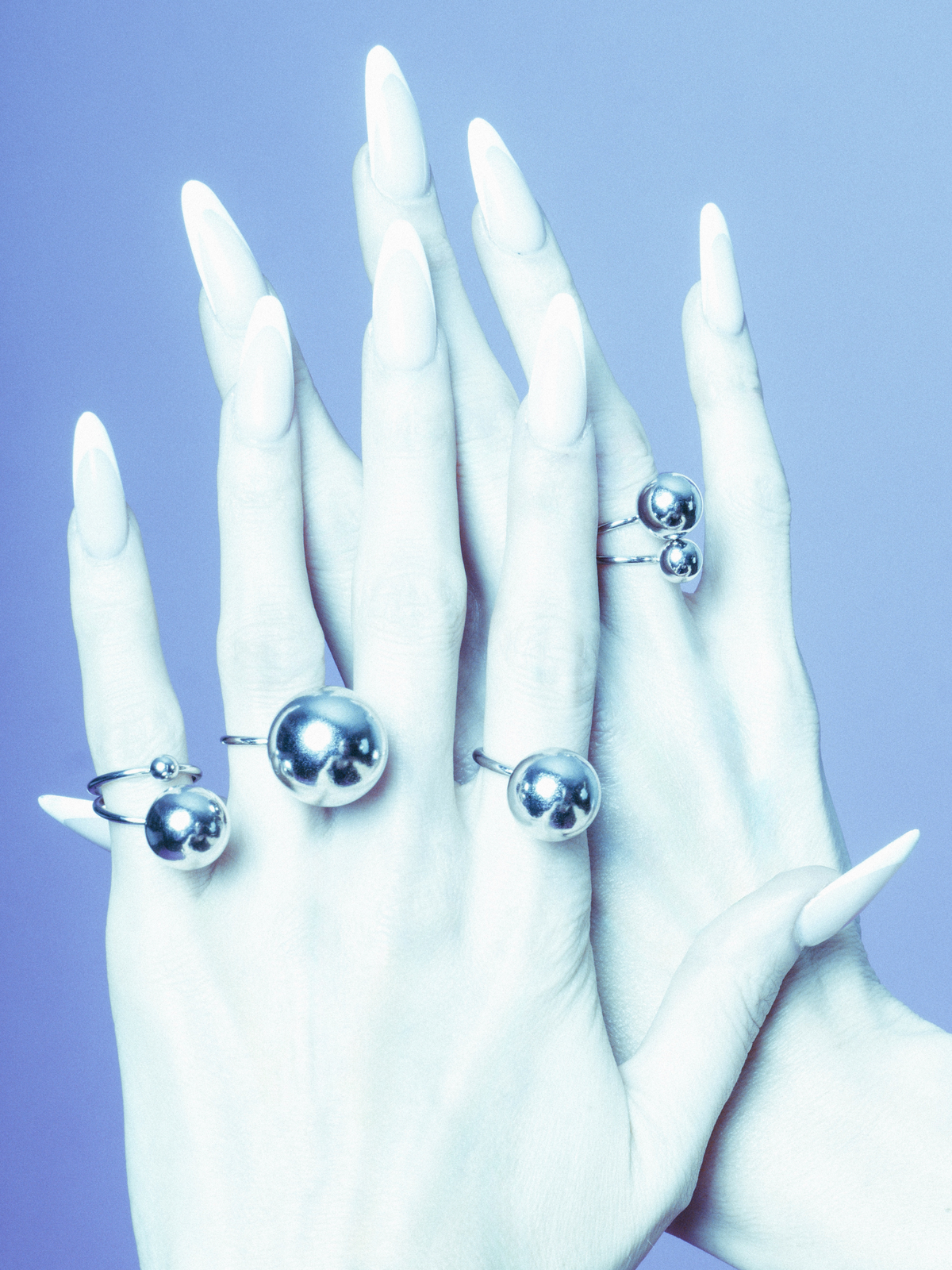 Long, pointed nails painted white wear the Orbita Silver Three Ring Stack by JAREDJAMIN Jewelry Online, featuring large spheres and a cool blue tone for a futuristic look. The fingers are artistically arranged to highlight the rings&#39; reflective surfaces.