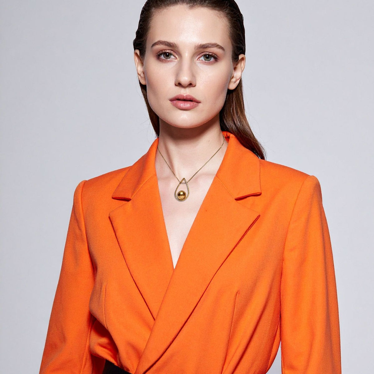 A woman with fair skin and straight brown hair wears an orange blazer, showcasing the Orbita Swing Gold Pendant Necklace by JAREDJAMIN Jewelry Online against a minimalist light gray background. She has neutral makeup and a modern look.