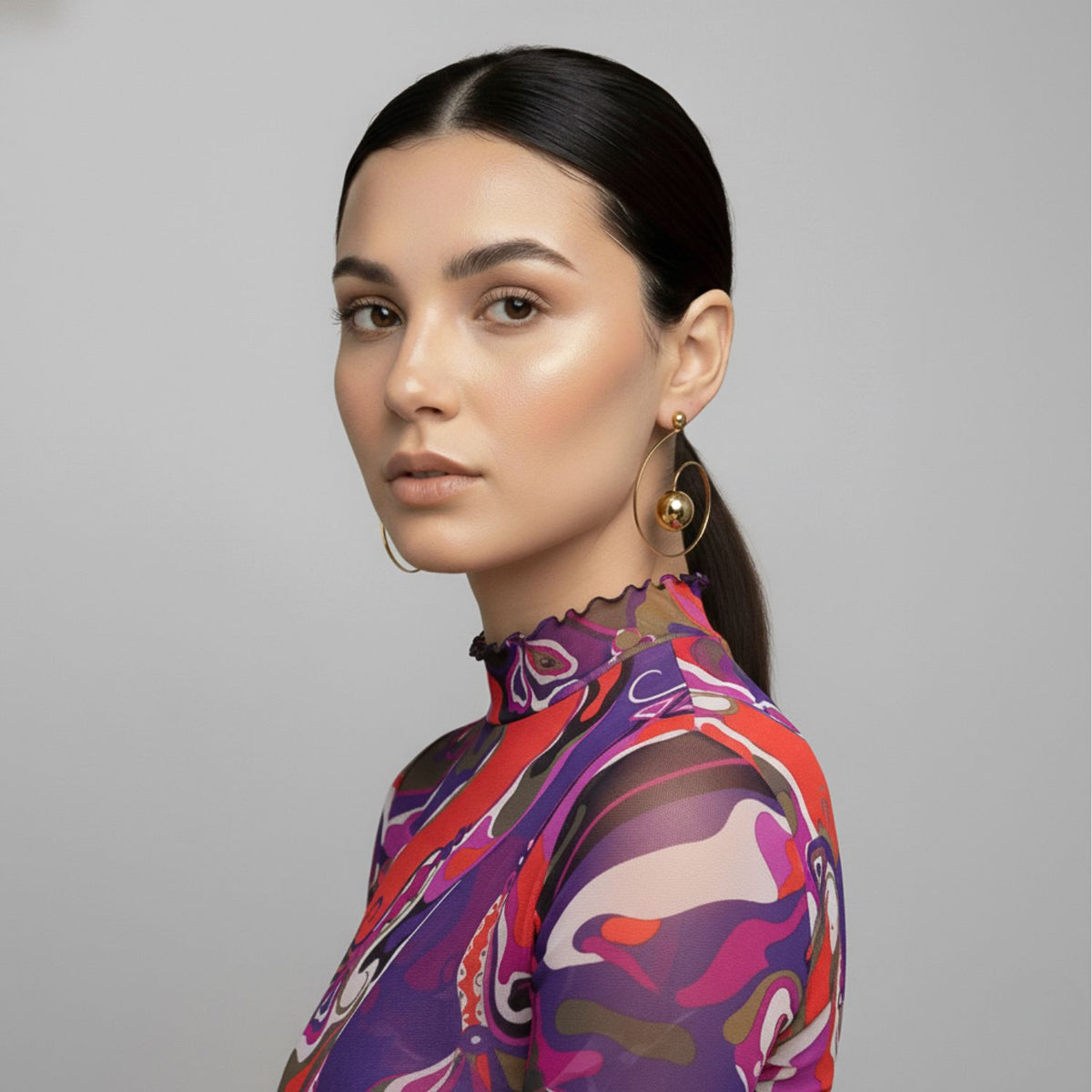 Un woman avec smooth, dark hair pulled back wears a sheer, high-neck colorful abstract top et JAREDJAMIN Bijoux en Ligne's Orbita Swirl créole Post boucles d'oreilles in 18K or. Her neutral makeup enhances her calm gaze against a gray background.