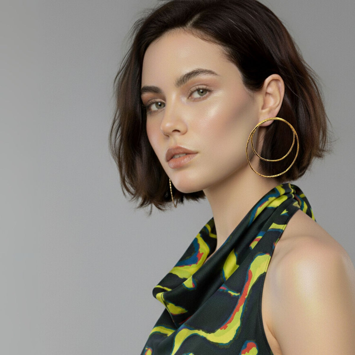A womanとshort brown hairとdewy skin looks to side, wearing Ottavia ゴールド Post イヤリング JAREDJAMINの ジュエリーオンラインとa sleeveless multicolored topとグリーン, yellow,とレッド accents against a plain light gray background.