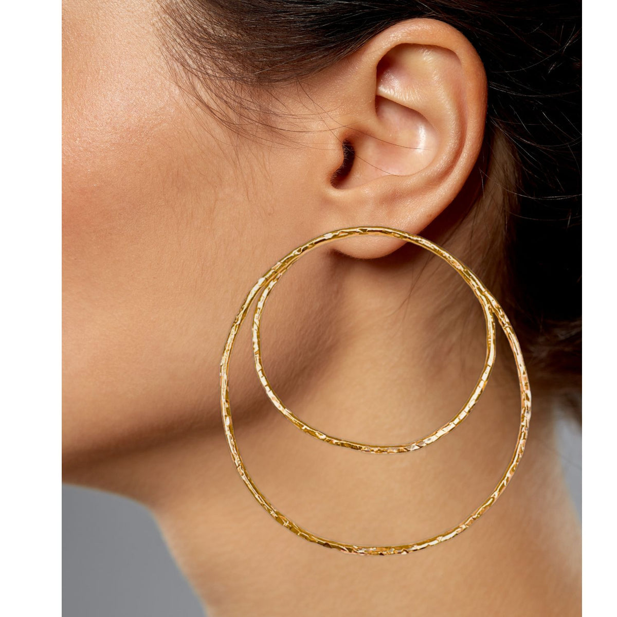 Close-up of someone wearing Ottavia Gold Post Earrings from JAREDJAMIN Jewelry Online. The 18K gold-plated textured brass hoops create a layered look on one ear, with a smaller hoop nested inside the larger. Light skin and partially pulled-back hair reveal this striking jewelry ensemble.