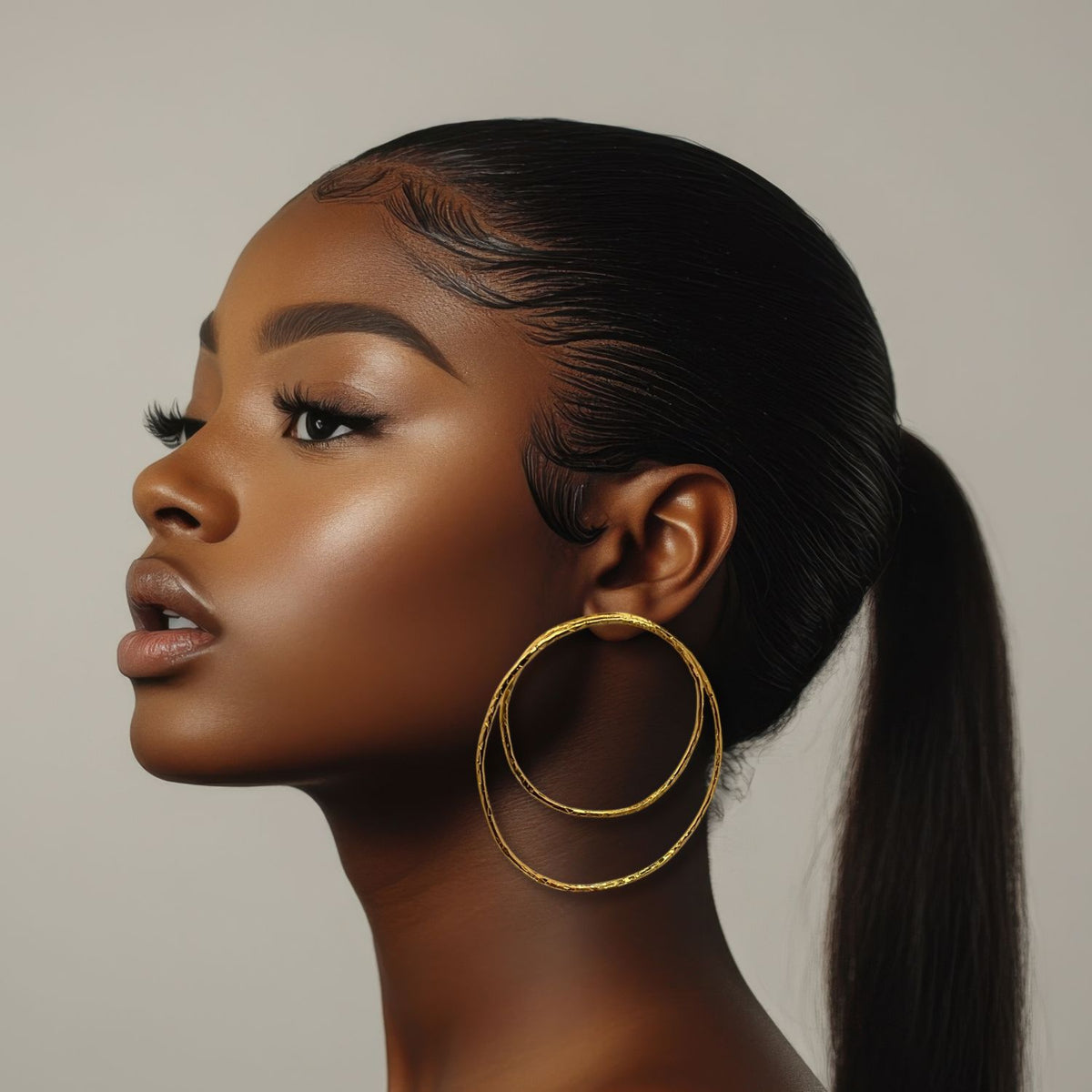 A young woman with smooth deep brown skin and sleek hair in a low ponytail models the Ottavia Gold Post Earrings by JAREDJAMIN Jewelry Online—large, textured, 18K gold-plated pieces—against a neutral background. Her makeup is flawless.
