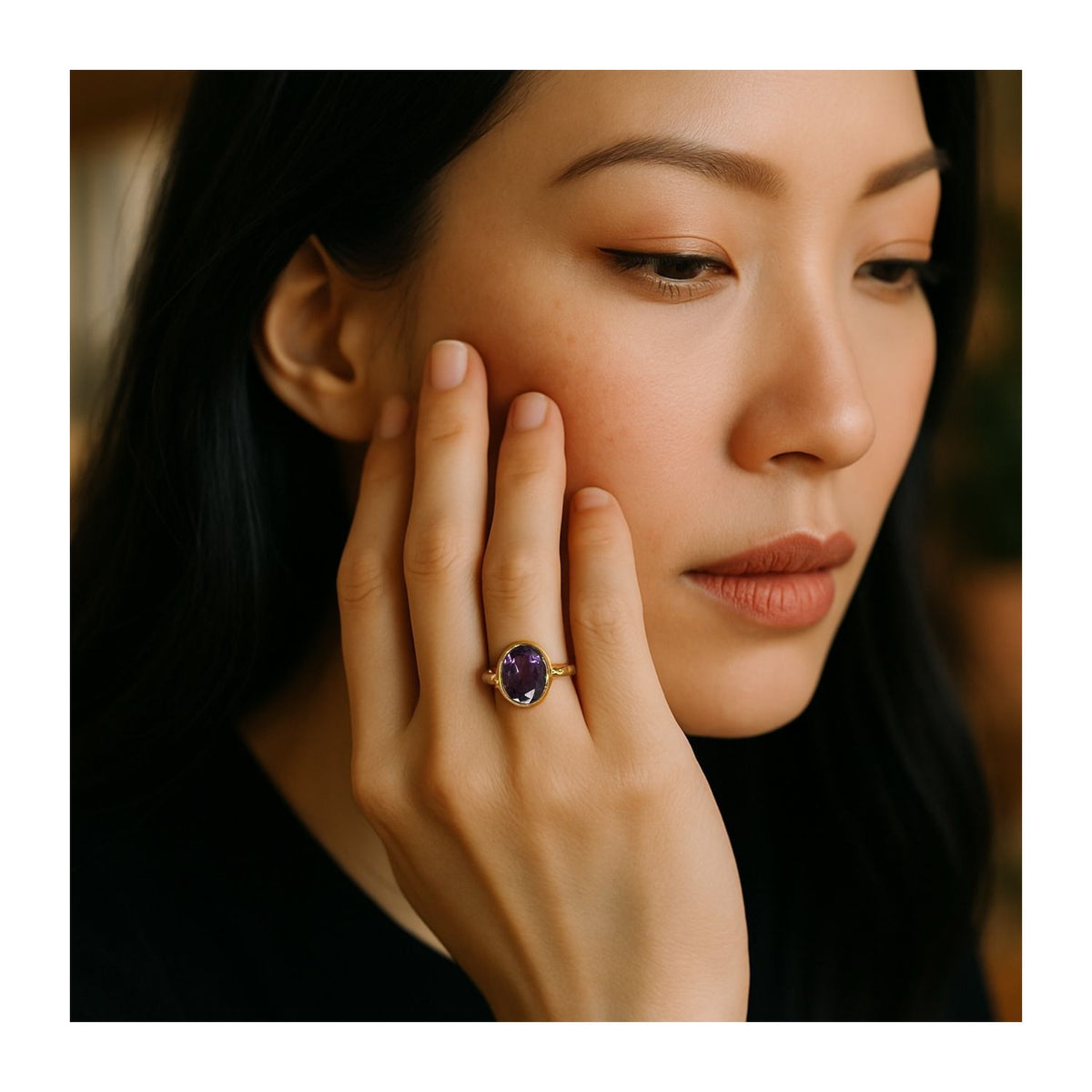 A woman with long dark hair gazes thoughtfully, her face resting on her hand. She showcases JAREDJAMIN Jewelry Online’s Oval Amethyst Gemstone Ring, an elegant 18K gold-plated statement piece. Her makeup is natural against a softly blurred background.