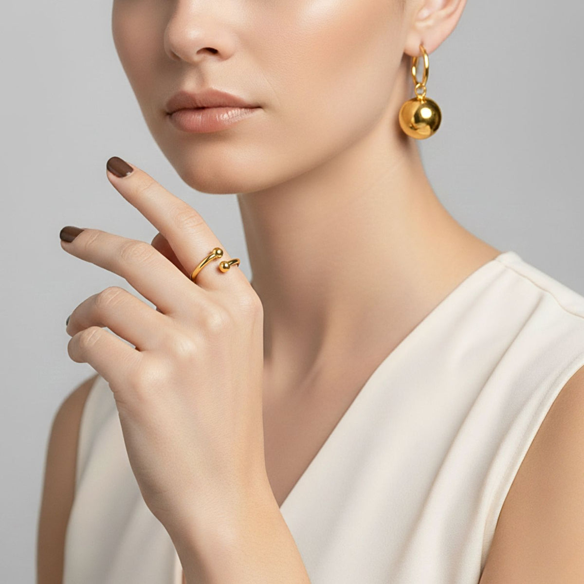 Un woman dans un sleeveless white top models or bijoux, avec a large créole boucle d'oreille avec a round breloque et the Over Easy Orbita bague par JAREDJAMIN Bijoux en Ligne. She has light skin, neutral makeup, short brown nails, et stands before a gray backdrop.