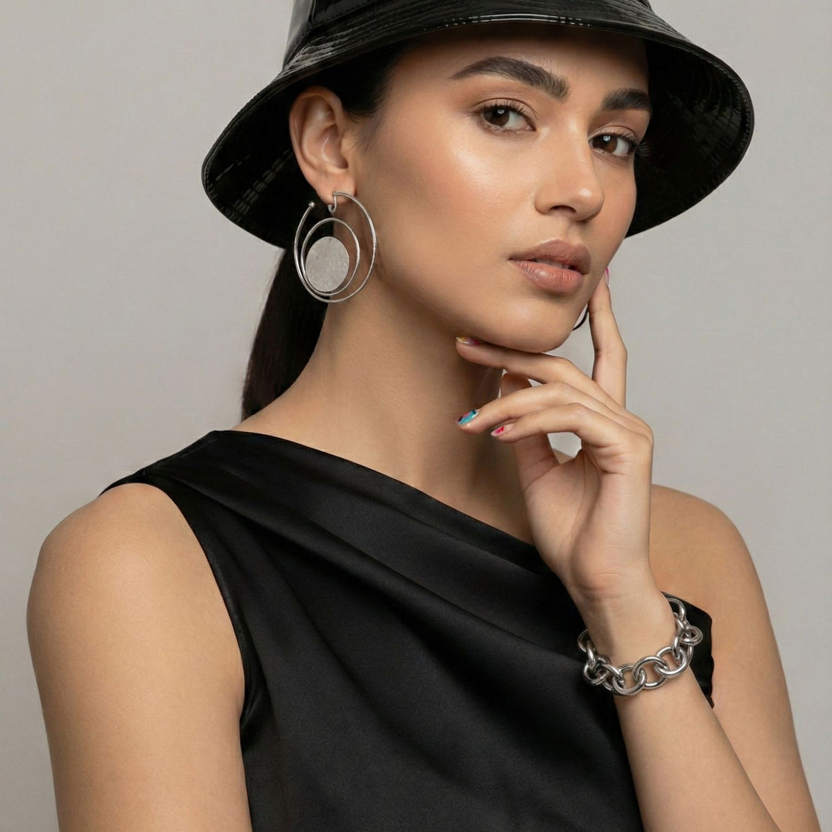 A woman in a black sleeveless top and shiny black bucket hat poses with her hand on her chin, wearing large silver hoop earrings and the Silver Parker Link Chain Bracelet by JAREDJAMIN Jewelry Online. She has subtle makeup and multicolored nails against a plain background.