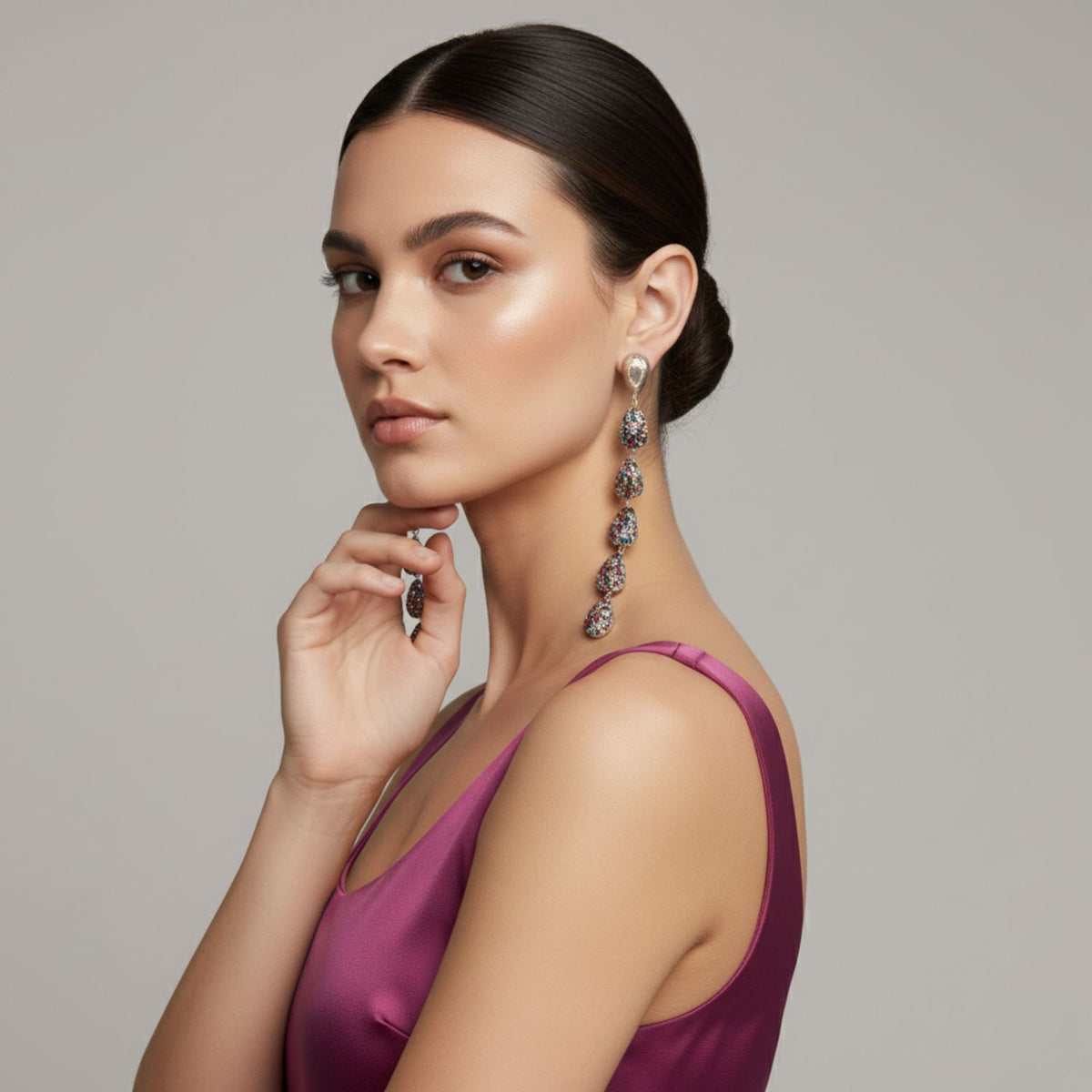 A woman with smooth, dark hair in a low bun wears a sleeveless magenta satin dress and JAREDJAMIN Jewelry Online's Pâques Drop Post Earrings. She poses in profile, chin on her left hand, gazing at the camera against a light gray background.
