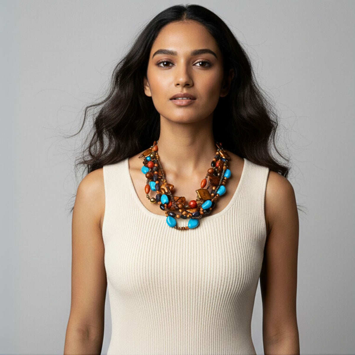 A womanとlong, wavy dark hair wears a sleeveless cream ribbed topと Passionate Sedona ネックレス JAREDJAMIN製 ジュエリーオンライン, 特徴とする Chalk ターコイズとアンバー beads. She stands confidently before a light grey background.