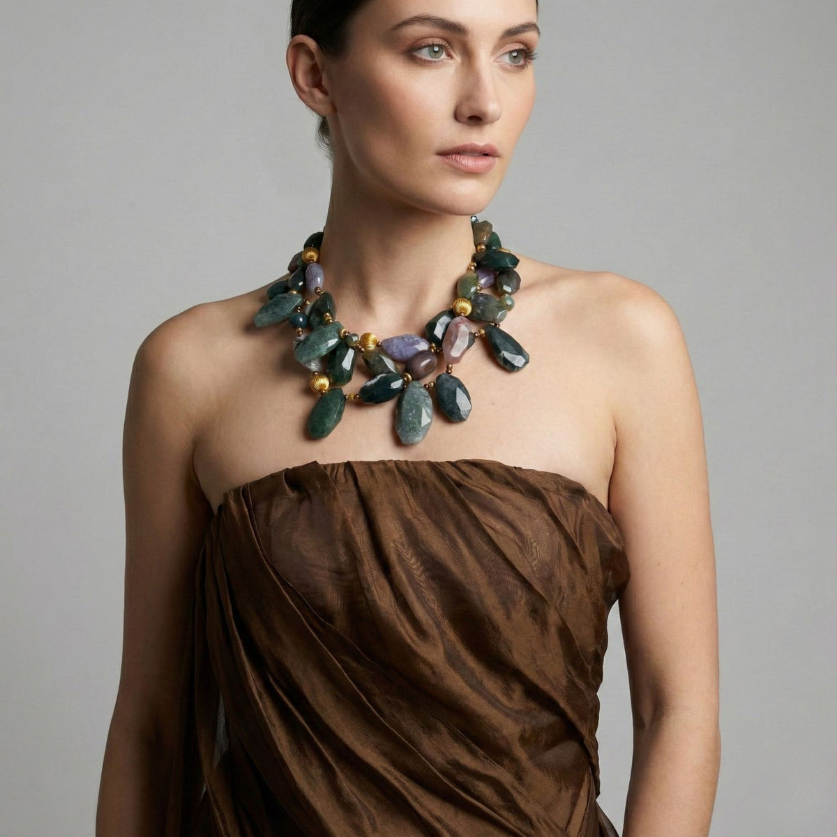 Un woman con light skin y dark hair pulled back poses en un strapless, texturizado brown dress against a gray background. She wears the Pebble Brook Jasper collar de JAREDJAMIN Joyería Online, con large multicolored stones y oro accents.