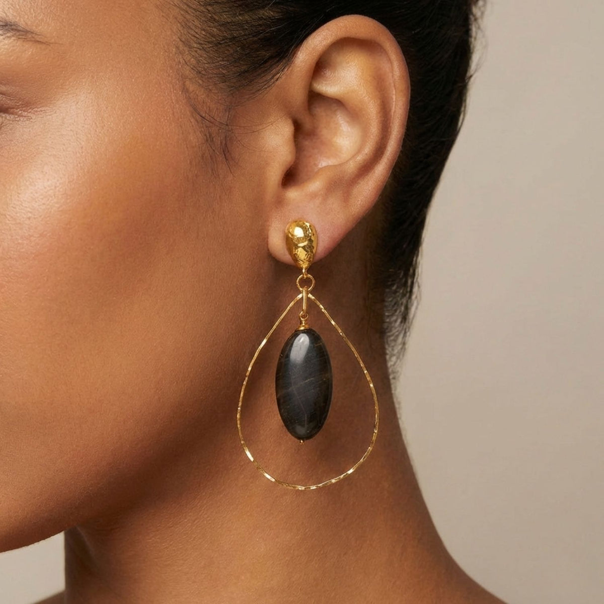クローズアップ of a woman's ear wearing JAREDJAMIN's Petra Jasper イヤリング—ゴールド plated 付きの small スタッド, ティアドロップ-shaped フープ,と大胆な ブラック stone centerpiece. Her hair is pulled backと background is neutral.
