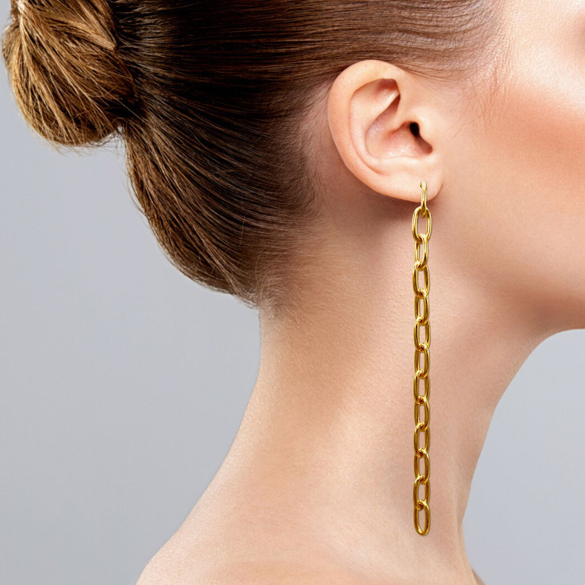 A woman profiles with an elegant bun, adorned with JAREDJAMIN&#39;s Chain Reaction Dangle Post Earrings. These striking 18K gold-plated earrings feature interlinked oval loops that nearly reach her shoulder, highlighted beautifully against a plain, soft gray background.