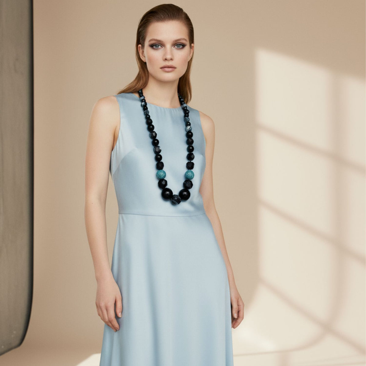 Un woman en un sleeveless, light blue dress stands en un softly lit room con beige walls y a large window casting shadows. She wears the Rock Of Gibraltar collar de JAREDJAMIN Joyería Online y has straight light brown hair con dramatic eye shadow.