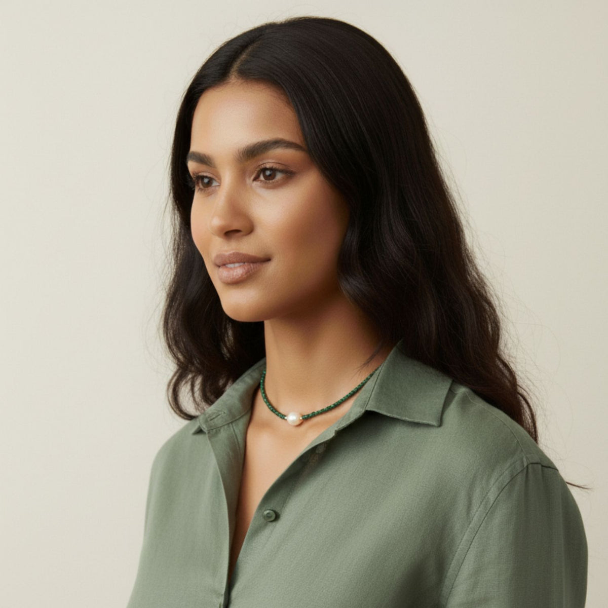 Un woman avec long, wavy dark hair wears a sage green collared shirt et the Rowan collier Malachite par JAREDJAMIN Bijoux en Ligne, avec freshwater perles et malachite beads. She looks calmly to her left against a plain beige background.