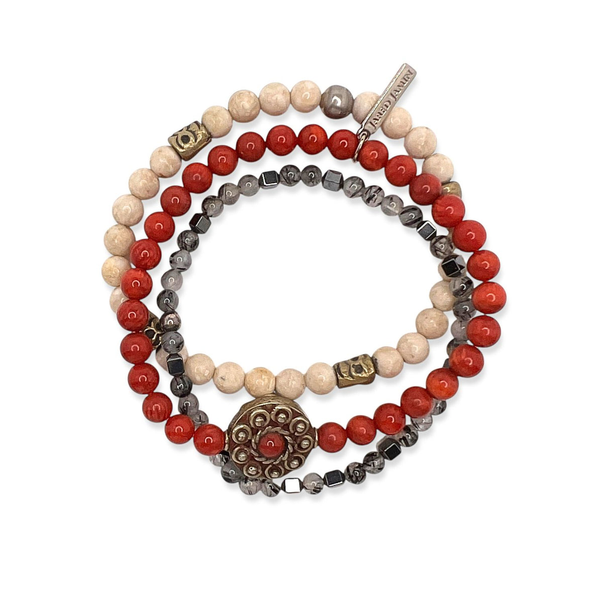 Los Red Barron pulsera Set de JAREDJAMIN Joyería Online features three coiled strands con cream, red, y metallic grey coral beads, a round metal dije con a red center, square brass accents, y a “Demure” tag.
